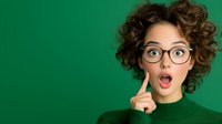 Young Caucasian woman with curly hair and glasses showing surprised expression with open mouth and finger on cheek against green background for advertising or blog content.