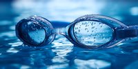 Swimming goggles with water drops floating on blue rippled water surface, dramatic close-up shot capturing reflection and transparency in competitive sports setting.