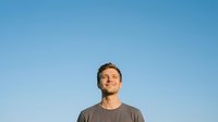 Male adult, Caucasian ethnicity, wearing casual gray t-shirt, looking upward with optimistic expression against clear blue sky background for lifestyle and motivational content.