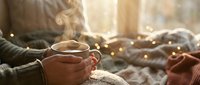 Cozy morning scene with hands holding steaming coffee mug in warm sunlight. Soft blankets and fairy lights create hygge atmosphere for relaxation and comfort.