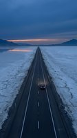 Straight asphalt road stretching across frozen salt flat at dusk with single vehicle driving toward distant mountains under twilight sky, conveying isolation and journey.