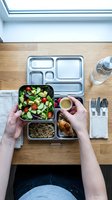 Overhead view of person holding compartmentalized meal prep container with fresh salad, grains, and protein on wooden desk with empty bento box and water bottle for lunch planning.