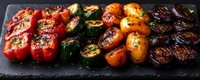 Grilled colorful mini sweet peppers and cherry tomatoes with herbs and spices arranged in rows on black slate board, showing charred, caramelized surfaces.