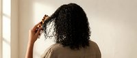 Young African American woman with curly hair applying serum or oil treatment to scalp in bright bathroom for healthy hair care routine.