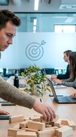 Caucasian male professional in beige sweater arranging wooden blocks at conference table in modern office with target symbol on glass wall, colleagues working in background, representing business strategy and teamwork.