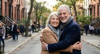 Happy senior Caucasian couple embracing on urban street with brownstone buildings. Elderly man and woman showing love and affection in city neighborhood setting.
