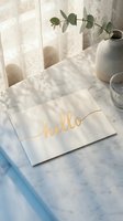 Elegant white envelope with gold foil hello script on marble surface, soft natural light, lace curtain, eucalyptus, and ceramic vase in background.