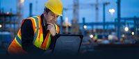 Construction worker in safety vest and hard hat using laptop computer at industrial site during evening hours with blurred city lights in background.