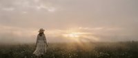 Woman in vintage white dress standing alone in misty cotton field at sunrise, atmospheric rural landscape with golden light breaking through fog.