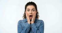 Young woman with curly hair showing shocked surprised expression holding hands on cheeks wearing denim shirt isolated on white background.