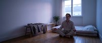 Young man practicing meditation in minimalist bedroom with natural light from window. Peaceful morning routine for mental wellness and mindfulness.
