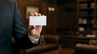 Professional man in dark suit holding blank white business card in office environment with wooden interior and blurred background for corporate branding.