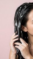 Young Asian woman applying hair mask treatment to wet dark hair against soft pink background for healthy haircare routine.