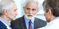 Senior businessman with white beard in gray suit discussing with colleagues during professional meeting in office environment.