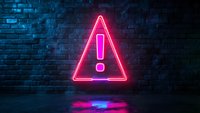 Glowing neon warning sign with exclamation mark on dark brick wall background with pink and blue light reflection on wet floor surface. Modern urban night scene.
