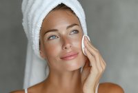 Young woman with white towel on head applying skincare product with cotton pad. Daily beauty routine, facial cleansing and moisturizing for healthy glowing skin.