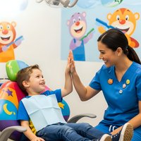 Young Hispanic female dentist giving high five to happy Caucasian boy patient in colorful pediatric dental office with cartoon characters on wall.