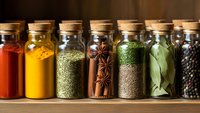 Row of glass spice jars with cork lids containing colorful herbs and spices on wooden shelf for cooking and culinary seasoning applications.