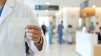 Doctor in white coat extending hand for handshake in modern hospital corridor with medical staff and blurred background, representing healthcare professional greeting and patient care.
