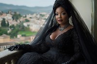 Elegant African American woman in black sequined dress and veil poses gracefully on balcony overlooking scenic Mediterranean hillside town landscape.