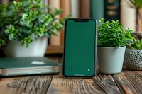 Modern smartphone with green screen display on wooden desk near laptop and houseplants, workspace mockup for app or website presentation.
