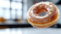 Freshly glazed vanilla donut with white icing floating in soft focus against blurred background, showcasing sweet pastry details and delicate sugar coating.