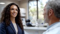 Young woman with long dark curly hair and medium skin tone wearing navy blazer, smiling during professional conversation with mature man in modern office setting with bright windows.