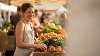 Young Caucasian woman shopping at farmers market with fresh organic produce and vegetables in background during sunny day.