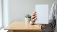 Businesswoman holding blank white card at wooden desk with plant, covering face for mockup and branding purposes.