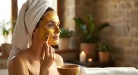 Young woman applying golden face mask in spa bathroom with towel on head enjoying skincare routine and wellness treatment at home.