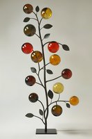 Decorative metal tree sculpture with amber and red glass spheres on branches with dark leaves, standing on black base against light background.