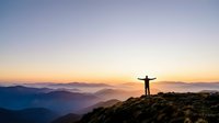 Person standing on mountain peak with arms raised celebrating success at sunrise with layered mountain ranges and golden sky horizon.