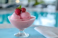 Pink raspberry ice cream scoops served in elegant glass bowl garnished with fresh berries and mint leaves against blurred pool background, summer dessert.