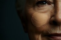 Close up portrait of senior Caucasian woman's face showing aging skin, wrinkles and foundation makeup application against dark background. Cosmetic and skincare concept.