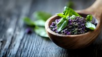 Organic purple basil seeds in wooden spoon with fresh green leaves on rustic table for healthy cooking and natural wellness supplements.