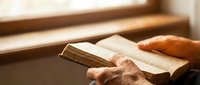 Elderly hands holding an open old book near a sunlit window, conveying wisdom, faith, and quiet contemplation in warm natural light.