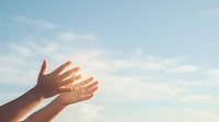 Human hands reaching toward bright sunlight against soft blue sky with white clouds creating inspirational moment of hope and spiritual connection.