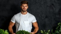 Young athletic male chef in white t-shirt holding fresh organic microgreens and herbs against dark textured wall in restaurant kitchen. Professional food preparation.