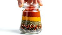 Hand opening glass jar filled with colorful layered spices including paprika, turmeric, herbs and peppercorns for cooking and seasoning on white background.