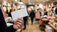 Person holding blank white card in retail clothing store with blurred shoppers and merchandise in background, suitable for promotional messaging and retail marketing campaigns.