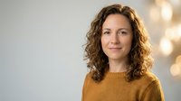 Professional middle-aged Caucasian woman with curly brown hair wearing mustard yellow sweater smiling confidently in modern office environment.