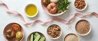 Overhead view of fresh tomatoes, cucumbers, nuts, grains, and olive oil arranged with pink ribbon on white surface for healthy cooking and nutrition content.