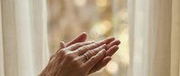 Senior woman with wrinkled hands praying or meditating by window with natural sunlight, expressing spirituality, faith, hope, and peaceful contemplation in home setting.