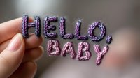 Female hand holding knitted purple text saying hello baby against blurred background, creative handmade lettering announcement for baby shower or birth celebration.
