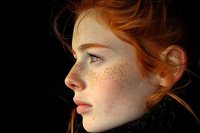 Side profile portrait of young redhead woman with natural freckles and glowing skin against dark background, wearing black turtleneck sweater. Soft dramatic lighting.