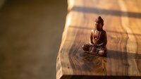 Small bronze Buddha statue sitting in meditation pose on wooden surface with warm sunlight and shadow patterns creating peaceful spiritual atmosphere.