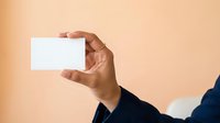 Close-up of male hand holding blank white business card against warm beige background for corporate branding and professional networking purposes.