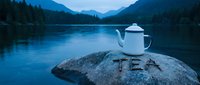 White enamel teapot on a lakeside rock with TEA written in stones, misty mountain forest backdrop at dusk, serene outdoor camping scene.