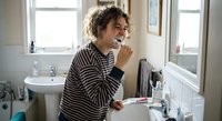 Young woman with curly hair brushing teeth in modern bathroom wearing striped pajamas. Morning dental hygiene routine and oral care habits at home.