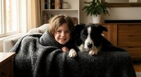 Young girl cuddling with border collie puppy under cozy knit blanket on living room sofa. Warm family bonding moment at home with pet companionship and comfort.